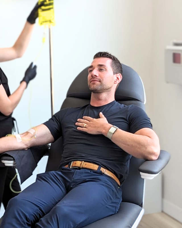 A man reclining in a medical examination chair with a vein visible on his arm, while a female healthcare worker in black scrubs stands nearby holding a yellow sign. The room has light walls and a framed abstract painting.